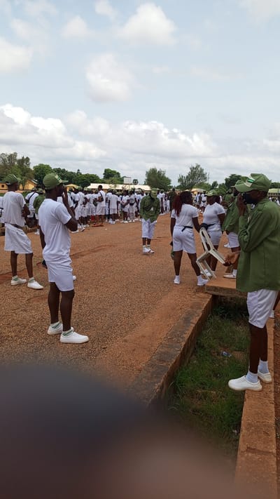The image depicts a group of young adults, most of whom are wearing white shorts and t-shirts with green caps, suggesting they are part of a uniform group, possibly a youth service or educational program. They are gathered outdoors on a gravel and dirt path, with some standing in rows and others milling about. In the background, there are buildings and trees, indicating an outdoor setting in a populated area. The sky is partly cloudy, suggesting daytime. The individuals appear to be engaged in some form of assembly or activity. The scene has the characteristics of an event taking place in a community or institutional setting, such as a school or training ground.
