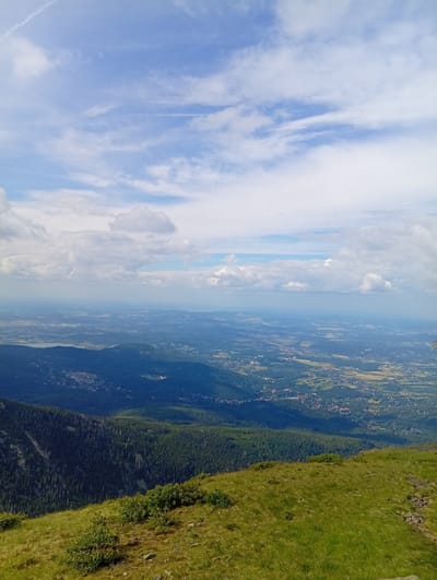 The image is a panoramic view from a mountain summit overlooking a valley and distant landscape. The foreground features a grassy slope with scattered rocks and low-lying green bushes. In the mid-ground, a dense, dark green forest covers the mountainside, with some lighter green patches and visible tree tops. Further into the valley, a scattering of buildings and settlements are visible, interspersed with fields of varying colors, suggesting agricultural activity. The background reveals a hazy, expansive landscape stretching to the horizon under a partly cloudy blue sky. The scene depicts a natural environment with signs of human habitation in the valley below. This view is likely from a location in or near Malá Úpa, Czechia, offering a vista of the surrounding countryside.
