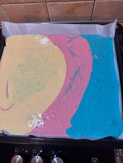 An overhead forensic image depicts a rectangular baking tray, lined with white parchment paper, containing a multi-colored liquid batter. The batter, approximately 2-3 centimeters deep, is primarily divided into three distinct sections: a pale yellow-cream hue on the left, a vibrant pink arc in the center, and a bright blue mass on the right. Visible swirling patterns indicate intermixing of the colors, with light blue streaks observed within the yellow and pink sections, and pink and yellow streaks within the adjacent colors. Numerous small air bubbles are present across the surface of the batter. The tray rests on the dark black surface of a kitchen appliance, likely an oven or stovetop, with three silver-and-black rotary control knobs partially visible along the bottom edge; a partial "Electrolux" logo is discernible near the rightmost knob. In the background, above the tray, a wall is visible, tiled with light brown or beige square tiles laid in a horizontal pattern. The scene is located in a kitchen within the city of Andratx, Spain.