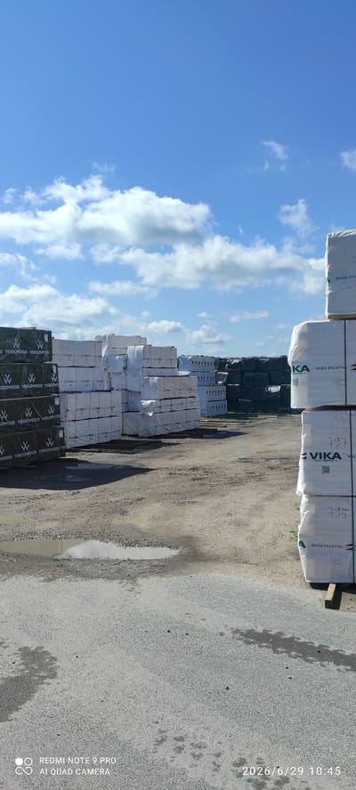 The image depicts an outdoor lumberyard in Verijärve, Estonia, under a bright blue sky with scattered clouds. The scene is dominated by large stacks of lumber wrapped in protective plastic sheeting. Some of the plastic coverings have brand names and text visible, such as "VERSOWOOD" and "VIKA MADE IN LATVIA". The ground is a mix of gravel and asphalt, with several puddles indicating recent rain. In the foreground, a watermark reads "REDMI NOTE 9 PRO AI QUAD CAMERA", and a timestamp "2026/6/29 10:45" is present. There are no people visible in the image. The overall impression is of a commercial outdoor storage facility for wood products.