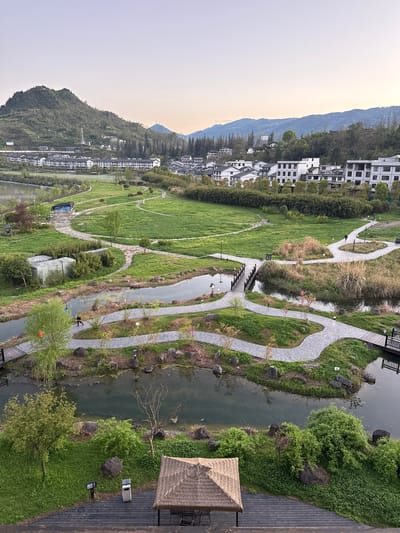 An elevated, oblique view captures a landscape in Zhuoshui, China. A waterway meanders through the scene, bordered by walking paths. The path is lined with greenery and decorative rock formations. Buildings cluster on the left, giving way to a grassy lawn dotted with trees and paths. Mountains are visible in the backdrop under a dusky sky. A thatched roof structure partially occupies the bottom right, adjacent to a wooden deck.