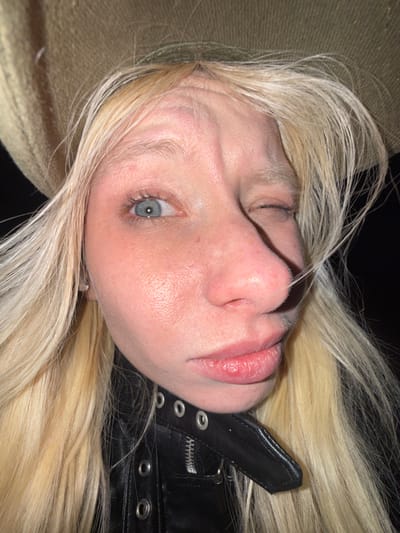 A close-up, rotated view captures a fair-skinned individual with platinum blonde hair. One blue eye is visible, partially closed. The subject's nose is prominent, and their lips appear full. They wear a black leather jacket with metallic eyelet details and a zipper. Part of a tan-colored, textured surface appears at the bottom left. The background is dark.