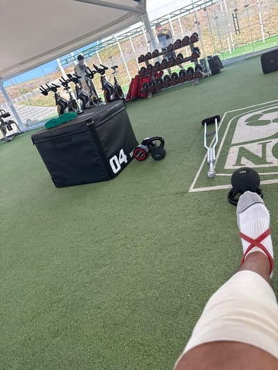 The image captures a first-person perspective, looking down at a person's leg and foot within what appears to be an outdoor or semi-outdoor gym facility.
In the immediate foreground, to the right, is a person's lower right leg and foot. The foot is clad in a white sock with grey stripes and accents. A red resistance band is wrapped around the ankle, crossing over the top of the foot. The lower leg, above the ankle, is encased in a light-colored, possibly beige or off-white, bandage or compression wrap. The person's skin tone is dark. The foot is positioned next to a black kettlebell, which prominently displays "16KG" in white lettering, indicating its weight.
The floor of the facility is covered in green artificial turf, resembling a sports field, with white markings forming part of a larger logo or text, where "S N" is visible.
Scattered across the artificial turf are various pieces of gym equipment. Near the person's foot, there is a silver or chrome piece of equipment, possibly a type of handle or part of a functional training apparatus, with a ribbed grip section. To the left of this, a black plyometric box (or jump box) is positioned, with the number "04" clearly marked in white on its side. Adjacent to the plyometric box are two smaller black weights, one of which shows "2.5KG" and "BRV" written on it.
In the middle ground and background, more gym equipment is arranged. Along the left side of the image, a rack of dumbbells with red handles and black weights is visible, ranging in size. Further behind the dumbbell rack, several stationary bikes or spin bikes are lined up.
The setting appears to be an open-air or partially enclosed gym, possibly under a large white tent or canopy structure, with support beams visible on the far left. Through glass panels or an open wall in the background, a view of an outdoor landscape can be seen, including what looks like a body of water (possibly a lake or ocean) and some buildings in the distance. The lighting suggests it is daytime, with bright natural light illuminating the area. In the upper left corner, a black bag or speaker with "JBL" written on it is partially visible.
The scene conveys an atmosphere of training or rehabilitation, given the specialized equipment and the bandage on the leg.