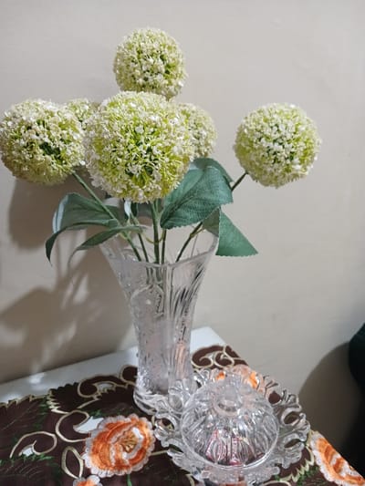 A clear glass vase, adorned with an embossed floral pattern, holds a bouquet of artificial white and pale green spherical flowers with green leaves. The vase rests on a dark brown embroidered placemat featuring orange and white floral designs. To the right of the vase, a small, clear, faceted glass dish with a lid sits on the placemat. The background is a plain, light-colored wall, with a shadow cast from the floral arrangement. The scene appears to be an indoor still life arrangement, likely on a table, in a residential setting in Geudonggeudong, Indonesia.