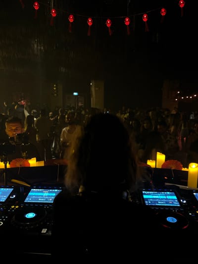 This is a dimly lit, vertical photograph taken at night, possibly outdoors or in a venue with an open-air feel, given the presence of what appears to be water in the foreground. The scene is dominated by a crowd of people, mostly silhouetted against a dark background. In the left foreground, a string of red lanterns hangs, their light illuminating the dark water below, creating ripples.
Central to the image, a DJ is visible, their head and shoulders in silhouette, facing away from the camera. They are positioned behind DJ equipment, including turntables and digital screens displaying waveforms and track information. One screen shows "03:24". To the right of the DJ, several large, bright yellow cylindrical lights, possibly candles or illuminated decorations, are prominent, casting a warm glow. Red paper umbrellas or decorative fans are also visible near the lights.
The crowd in the background appears to be enjoying themselves, with some individuals leaning in and gesturing. The overall atmosphere suggests a lively event, possibly a music performance or party. The time of day is clearly night, and while no specific weather is indicated, the presence of water suggests a potentially humid environment. The red lanterns and paper umbrellas hint at an Asian cultural influence, fitting with the provided location context of Ban Bang Thao, Thailand. The emotions conveyed are generally positive and energetic, driven by the music and the gathering.