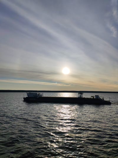 A barge is seen silhouetted against a bright sun setting over a body of water. The barge is long and low, with structures at both ends. The water is rippled and reflects the sunlight. A dark treeline is visible on the horizon. The sky is streaked with pale clouds. The location is likely Shchetinskoe, Russia.