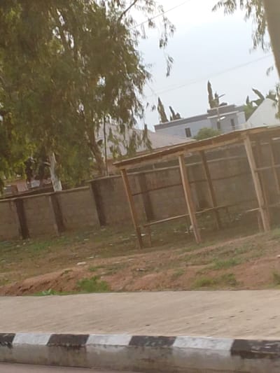 The image shows an outdoor scene in Kporva, Nigeria, featuring a wooden structure with a corrugated metal roof, possibly a shelter or stall. The structure is open on at least two sides and has horizontal wooden supports. Behind the structure, a low, rough concrete wall runs horizontally across the frame. Beyond the wall, a multi-story building with grey and white facades is visible, along with several tall, slender trees. The foreground of the image shows a paved surface with a black and white striped border, suggesting a road or pathway. The overall environment appears somewhat dry with sparse green vegetation and brown earth. The lighting suggests it may be late afternoon or early morning, with a bright, overcast sky. There are no people or animals visible in the image.