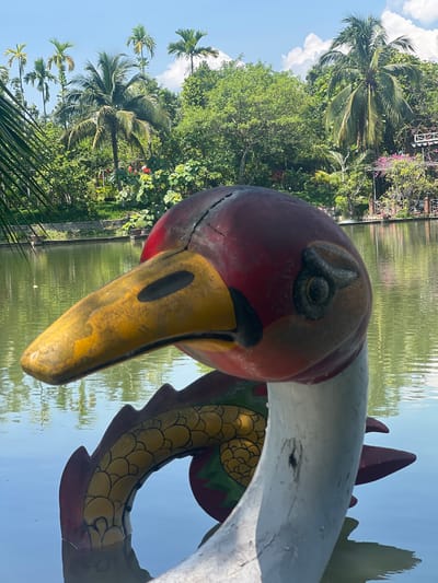 A brightly colored, weathered statue of a mythical creature, possibly a dragon or phoenix, floats in a tranquil body of water. The statue's head, predominantly red with a yellow beak and black eye details, is in the foreground, slightly off-center to the right. Its body, adorned with yellow scales and red accents, curves into the water. The background features a lush, green landscape with numerous palm trees, dense foliage, and a clear blue sky with scattered white clouds. The water reflects the surrounding greenery and sky. The scene is likely a public park or recreational area within the city of Suối Tân, Vietnam, given the characteristic tropical vegetation.