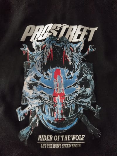 This image is a close-up, high-resolution photograph of a black textile surface, likely clothing, featuring a graphic print. The item is located in Linkebeek, Belgium. The print is centered and prominent, displaying several distinct elements:

At the top, the word "PROSTREET" is rendered in stylized, fragmented white capital letters with a distressed appearance.

Below this text is a complex graphic dominated by a menacing, stylized wolf-like head or a creature resembling an armored helmet. This central image is depicted primarily in shades of blue, white, and black, with notable red accents. Sharp fangs are visible at the mouth, from which red, blood-like drips extend downwards. Similar red streaks are present across the face and eyes of the creature. The design incorporates mechanical or skeletal elements, suggesting a fusion of organic and inorganic forms.

Below the main graphic, two lines of white text are present:
The first line reads "RIDER OF THE WOLF" in a bold, sans-serif font.
The second line, in a smaller font, reads "LET THE HUNT SPEED BEGIN".

The black fabric background shows a subtle woven texture, and the print exhibits slight variations in color saturation and detail consistent with screen printing on textile. No surrounding environment or human presence is visible in the frame.