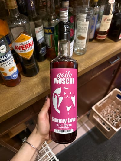 The image shows a person holding a bottle of "Gummy-Love" liquor with a pink label featuring a cartoon cat. The label also includes "geile MUSCHI" text. The bottle is surrounded by other liquor bottles in what appears to be a bar or storage area. Several brands such as "Averna", "Mazzotto", and "Kümmel" are partially visible. The liquor is produced by Vision Spirits GmbH, and the bottle states they are located at Neudenburger Straße 21, D-28207 Bremen, Germany. No location of Bruchköbel, Germany is mentioned on the bottle. The person holding the bottle has a bracelet and manicured nails.