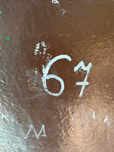 A close-up shot of a brown, seemingly painted, surface, showing signs of wear and tear. White markings are present on the surface, including a large "6" or "9" shape, a symbol resembling a stylized "R" with a cross, and a mark that looks like a capital "V" or inverted "A". The precise nature and meaning of the markings are unclear. The setting is Sayansk, Russia, though this can not be confirmed by the image provided. There are no people or interactions visible. The object depicted is simply a marked surface of unknown composition.