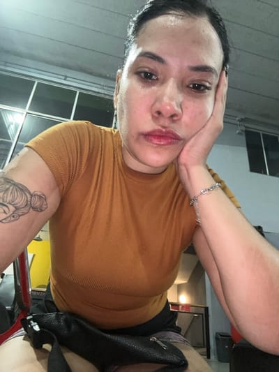 An adult female, with dark hair pulled back and a visible sheen on her face indicative of perspiration, is captured in a close-up, chest-up view. She wears an orange/rust-colored ribbed short-sleeved top and dark shorts or a skirt. Her left hand supports her chin, conveying a pensive or weary expression as she looks directly at the camera. A stylized tattoo of a head with hair in buns is visible on her left upper arm. A silver-colored chain link bracelet adorns her right wrist. A black fanny pack with a silver zipper rests on her lap. The indoor environment features a light-colored, paneled ceiling. In the background to the left, a multi-paneled window or glass partition reflects light, and parts of red and black exercise equipment with a yellow accent are visible. The background to the right is dimly lit, showing a light source and a dark object, possibly a waste bin or gym accessory. The scene is located in Ciudad del Libertador General San Martín, Argentina.