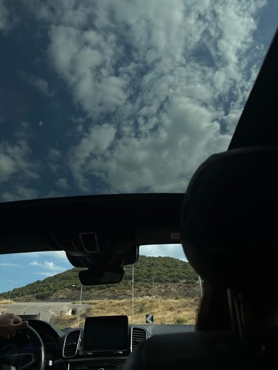 The image is a first-person perspective from inside a car, looking through the sunroof and windshield. The sky above is a mix of deep blue and scattered white clouds. Through the windshield, a green, tree-covered hillside dominates the background, with a winding road visible in the middle ground. The foreground shows the interior of the car, including a steering wheel, dashboard with a touchscreen display, and the back of a headrest. A hand is visible on the steering wheel. The scene appears to be a scenic drive on a road likely near Agios Andreas, Greece, given the mountainous terrain and vegetation.