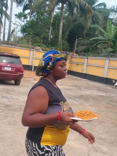 An adult female is depicted in profile, walking or standing outdoors in Itu, Nigeria. She has dark skin and is looking towards the right of the frame. Her head is covered by a blue, yellow, and black patterned fabric wrap. She wears a black sleeveless top with "LAKERS" in purple and yellow, a yellow fabric wrapped around her midsection, and black and white zebra-print patterned pants. Red bracelets are visible on both wrists. She holds a white rectangular plate in her left hand, containing a mound of orange-brown granular food, consistent with jollof rice. In the background to the left, a red SUV with license plate "ZLL 84EJ" is parked. To the right, a yellow concrete wall topped with barbed wire runs horizontally. The overall background features numerous tall palm trees and dense green foliage, including banana plants. The ground is a light-colored paved or concrete surface.