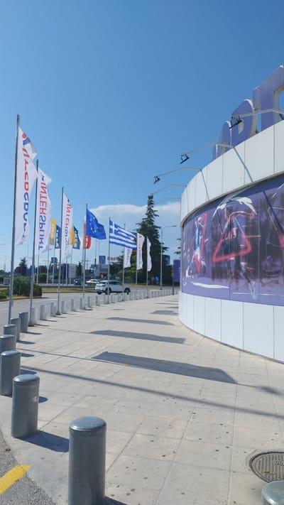 A frontal, medium shot captures a modern building with a curved facade adorned with a large purple and blue mural. The building is situated in Pylaia Municipal Unit, Greece. In front of the building, a row of cylindrical concrete bollards lines a paved walkway. A line of flags, including the European Union and Greek flags, flutters on poles along the walkway. Behind the flags, a white car is visible on a road. The sky above is clear and blue, with a few scattered clouds. The scene is illuminated by bright sunlight, casting long shadows from the bollards and flags onto the pavement.