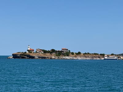 The image shows a panoramic view of the coastline near Atiya, Bulgaria. A small island with a lighthouse, several buildings, and sparse vegetation is prominent in the center left of the frame. The island is rocky and slopes down to the sea. The sea itself is a deep blue, with a gentle ripple on its surface. In the distance, on the mainland, a few more buildings and trees can be seen. A modern ferryboat is docked on the right side of the image, suggesting a port or harbor area. The sky is clear and a bright, uniform blue. There are no people visible in the image. The scene conveys a sense of calm and tranquility, with a focus on the natural landscape and man-made structures integrated into it.
