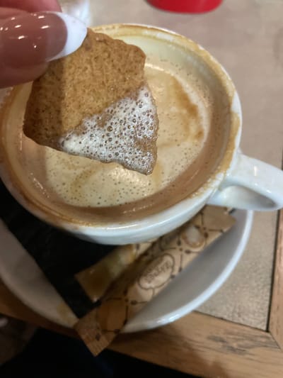 The image presents a close-up, high-angle view of a person dipping a biscuit into a frothy hot beverage. A hand, adorned with glossy, French-manicured nails, holds a light brown, rectangular biscuit, the lower part of which is submerged in the drink. The biscuit's submerged portion is coated with small bubbles from the beverage's foam.
The drink, likely coffee or a similar milky beverage, fills a white ceramic mug, which features a distinct handle. The mug sits on a matching white saucer. Resting on the saucer beside the mug are two brown sugar packets. The word "ZORBA" is clearly visible on one of the sugar packets, indicating a brand.
The setting appears to be indoors, possibly a cafe or restaurant in Chaidari, Greece, suggested by the arrangement of the coffee, biscuit, and sugar. Hints of a wooden surface, possibly a table, are visible in the background, along with other blurred elements. The scene evokes a moment of quiet enjoyment, focusing on the simple pleasure of a hot drink and a biscuit. The time of day is not explicitly shown but is consistent with a coffee break.