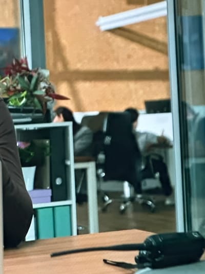 The image shows an indoor scene that appears to be an office. In the foreground, a wooden desk surface is visible with a black object, likely a walkie-talkie, lying on it. Behind the desk, there is a shelving unit with various items, including a white vase with a plant, a purple box, and two teal boxes. Further in the background, through a glass partition, two individuals are partially visible. One person is seated in a black office chair, leaning back with their arm extended, and appears to be wearing a light-colored shirt. Another person is seated behind them, also in a chair, with their head slightly obscured. The wall in the background has an orange, textured finish. There is a white fluorescent light fixture mounted on the ceiling. The scene is underexposed and slightly out of focus. The location is identified as Dondușeni, Moldova, suggesting this is an office space within that city.