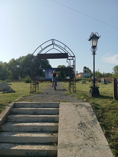 The image shows a bright, sunny day in Новокопылово, Russia. In the foreground, there are concrete stairs leading up to a gravel path. To the right of the stairs is a concrete ramp. Beyond the path is an ornate, wrought-iron archway with Cyrillic lettering that appears to say "ВЛАДИМИРСКОЕ". A young person is riding a bicycle through the archway. To the right of the archway is a tall, decorative lamppost. The background features a grassy field, trees, and a brick building. A banner with Russian text and a bear emblem is visible behind the archway. The sky is clear and blue.
