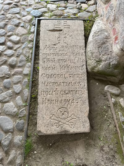 This image shows a weathered stone slab with carved inscriptions and a skull and crossbones motif, positioned on a cobblestone path. The slab is framed by a metal border. The inscription appears to be in Cyrillic script. The setting suggests an outdoor historical or memorial site, possibly a cemetery or religious complex. Given the context of the "Solovetskiy Stone" from Solovetskiy, Russia, this is likely a grave marker or memorial to victims of repression. The stones and surrounding earth show signs of age and exposure to the elements, with some moss and grass growing in crevices. A large, rough-hewn stone and a section of brick wall are visible to the right of the slab, further indicating an ancient or historical location.