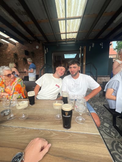 This image captures a daytime scene at an outdoor or semi-enclosed patio area of an establishment in Hornsea, United Kingdom. The foreground features a light-colored wooden plank table, upon which a person's right hand, adorned with a large, dark-faced wristwatch with a brown strap, is resting. The table holds several drinking vessels: two glasses containing dark stout (one a branded Guinness glass), two stemmed cocktail glasses with light-colored foamy drinks, multiple empty or partially empty clear glasses, and a glass with a straw. A small metallic ashtray is also present.
In the midground, three individuals are seated at the table. On the left, an elderly female with gray hair and sunglasses wears a multi-colored, abstract-patterned shirt and is looking to her left. Next to her, a young male in a white t-shirt with a red "Kellan" logo leans slightly to his right, smiling at the camera. To his right, another young male with dark curly hair, wearing a white t-shirt with "Forever Faster" text, looks directly at the camera. A fourth person is partially visible seated to the right.
The background reveals the establishment's structure: a brick wall with hanging plants on the left, and a dark teal-colored wall with an open doorway leading indoors on the right. Additional patrons are visible in the background: a blonde female and an older male in a striped shirt are standing on the left, while another blonde individual is partially visible on the far right. The patio is covered by a roof constructed with wooden beams and corrugated translucent panels, allowing natural light to penetrate. String lights are strung along the roof structure. The floor is covered with patterned tiles.