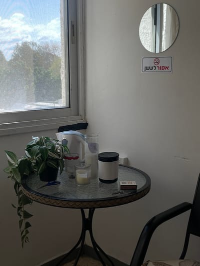 The image depicts an indoor corner space within a building in the Brener Regional Council, Israel. A round, glass-topped table with a woven-textured rim and black metal legs occupies the lower-center and lower-right foreground.
On the table, a black potted plant with trailing variegated green and white leaves is positioned on the left. To its right are a white electric kettle, a small red and white container, a tall clear glass cylinder, and a clear glass votive holding a white candle. Further to the right, a black and white cylindrical container with a black lid is present, alongside a small brown matchbox and a metallic object resembling a key. A pair of blue scissors is partially visible beneath the plant pot. The rightmost portion of a black chair with a patterned cushion is visible in the lower right.
The wall behind the table is light-colored, showing minor surface imperfections. An electrical outlet is present on the wall, approximately centered above the table. Above the outlet, a white rectangular sign displays a red circle-slash "no smoking" pictogram and the Hebrew text "אסור לעשן" (No Smoking). Above this sign, a circular mirror is mounted, reflecting a window frame.
To the left, a large window with a white frame and visible screen pane offers a view of the exterior. Black tape is affixed to the lower section of the window frame near the corner. Beyond the window, a bright blue sky with white clouds is visible above dense green trees and foliage.