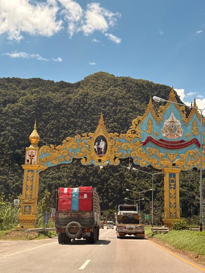 The image depicts a road leading towards a large, ornate archway with intricate gold and turquoise designs, likely marking an entrance or border. The archway features a crest at its apex and images of people on either side. Behind the archway, a lush, tree-covered mountain rises steeply under a bright blue sky with scattered white clouds. Two trucks are visible on the road, one in the foreground with a red and blue tarp covering its cargo and another in the middle distance. The truck in the foreground has a spare tire mounted on the back. Streetlights are present on either side of the road, and some vegetation lines the roadside. The overall scene suggests a location in Phang-nga, Thailand, characterized by natural beauty and cultural architectural elements.