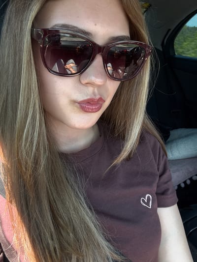A woman with blond hair is lying down in a car. She is wearing sunglasses and a brown shirt with a small white heart on the chest. The car interior is visible, including parts of the dashboard, seat, and a headrest. The reflection in her sunglasses shows details of a building and other objects. There is no specific information available to place the location in Levski, Bulgaria.