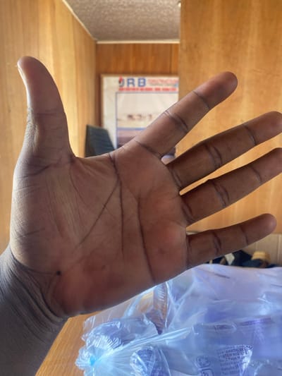 The image displays the open left hand of an individual, palm facing the camera, dominating the foreground. The skin is dark-toned, revealing prominent palm creases and several small, dark spots, including a distinct one on the thenar eminence near the wrist.
The background is an indoor environment, largely out of focus. Wooden paneling covers the walls. On the far wall, a white poster or sign is visible, bearing the red text "DRB" at its top, followed by blurred black text. To the left of the poster, a dark, possibly upholstered object is partially visible.
In the lower right foreground, beneath the hand, a large, translucent plastic bag filled with numerous smaller, clear plastic items is present on a wooden surface. Text printed on this bag identifies "PRODUCED & PACKED SYSTEMATIC SERV SYSTEM NIG. LTD." and provides an address: "No 7 Faga Mains Kofan Kona, Kaduna State," along with a partial phone number.
The location is an indoor setting in Kaduna State, Nigeria, specifically referenced to the Kofan Kona area of Zaria, within which Gyellesu is also situated.