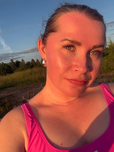 A woman with fair skin and green eyes is taking a selfie in Sapunov Bor, Russia, during sunset. She has her hair pulled back and is wearing a pink tank top and a silver hoop earring. The setting sun casts a warm, orange glow on her face and shoulders. In the background, there are trees and tall grass, with a cloudy blue sky above.