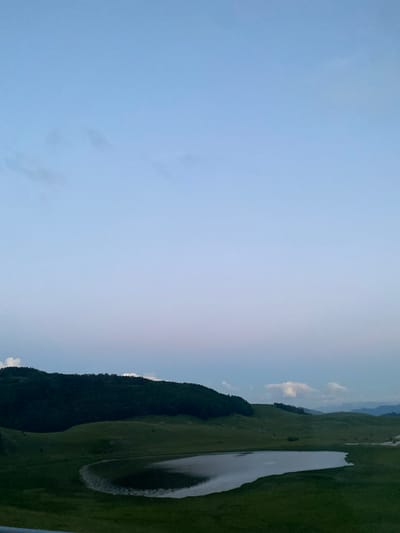 An outdoor, high-angle shot shows a body of water resembling a lake or pond in the foreground, reflecting the pale blue sky and some scattered clouds. Surrounding the water are rolling green hills covered in grass, with a dark, densely forested hill forming the backdrop. The sky is a gradient of light blue, fading to a lighter hue near the horizon, with a few wispy clouds. The scene is serene and natural, with no people or man-made structures visible apart from what might be a slightly paved or rocky edge along the lake. The image appears to be taken during twilight or early morning due to the soft lighting and the muted colors of the sky. The location is identified as Pašina Voda, Montenegro.