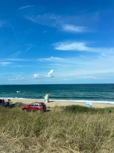 This is a photograph of the Black Sea coast near Dyuni, Bulgaria. In the foreground, dry, tall grass lines a dune overlooking the beach. A red SUV is parked on the edge of the dune, facing the water. On the sandy beach, there are a few beach umbrellas, chairs, and what appears to be camping gear. A colorful, inflatable advertising stand is also visible on the beach, and a blue and white sign is partially obscured by the grass. The sea is a deep turquoise color with moderate waves crashing on the shore. The sky is a vibrant blue with scattered white clouds. The overall scene depicts a relaxed, coastal setting.