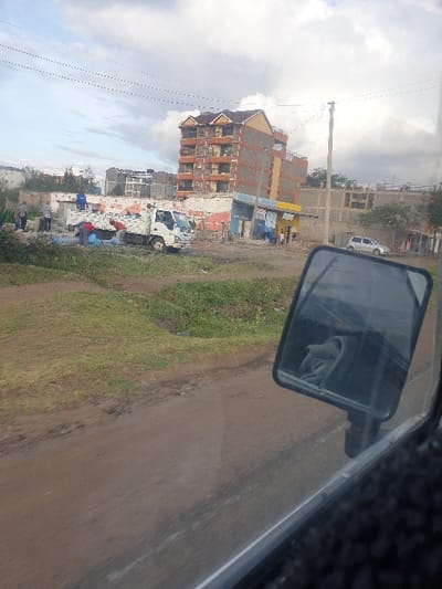 The image depicts a semi-urban scene in Mumbuni North ward, Kenya, viewed from inside a vehicle. The sky is overcast with heavy clouds, suggesting a cloudy day.
On the left side of the frame, a prominent multi-story building stands, featuring a brown gabled roof and exposed brick or concrete block construction. Several smaller, less distinct structures are visible around and behind it. A white utility pole with multiple power lines runs vertically through the middle ground, extending towards the left. Below the main building, a white truck is parked, and further down the road, another white car can be seen.
The foreground and right side of the image show a wide, unpaved dirt road with a grassy verge on its immediate left. The road appears somewhat uneven and dusty. In the upper right corner, the vehicle's side-view mirror is visible, reflecting the partial profile of a person's face, likely the driver or a passenger.
Several indistinct figures are scattered near the buildings on the left, with one person wearing a red top being slightly more discernible among them, suggesting daily activity or possibly work. The overall setting indicates a developing area, characterized by its architecture and unpaved roads. No legible text is visible in the image.