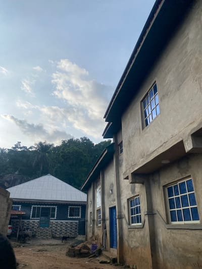 This image depicts an outdoor scene in Igarra, Nigeria, featuring two primary buildings under a partly cloudy sky. On the right, a multi-story building constructed from unfinished light grey concrete or stucco dominates the foreground. It features multiple windows with white frames and reflective blue glass, arranged in a grid pattern, and a prominent dark roof overhang. A blue door is visible on its lower level. To the left and slightly further back, a second building with a distinctive pyramidal corrugated metal roof is present. Its facade is dark, accented by a patterned tile wainscoting on the lower half and white decorative trim. The ground in the foreground consists of dirt, with visible piles of earth and construction materials. A dark animal, consistent with a dog or goat, stands near the base of the left building. In the background, a dense cluster of green trees, including palm trees, ascends a hillside, with visible rock formations at its base. The sky is light blue with scattered white and grey clouds, indicating daylight conditions. No human figures are visible.