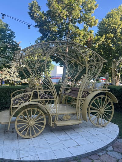 A decorative, ornate yellow carriage sculpture is displayed outdoors on a tiled circular platform. The carriage features intricate metalwork, including decorative scrolls and a lattice roof. It has wooden seats and large spoked wheels, resembling an antique carriage. In the background, lush green trees and bushes are visible, with a clear blue sky above. String lights are hung between trees, suggesting a park or outdoor event space. The environment appears to be a well-maintained public area, possibly a park or plaza within the city of Dubăsari town council, Moldova. There are no people visible in the image.