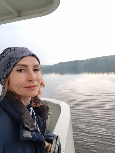 A woman wearing a grey patterned bandana and a dark blue quilted jacket is taking a selfie on a boat. Her head is turned slightly to her left, and she is looking at the camera with light makeup on. She has auburn hair peeking out from under her bandana. In the background, there is a calm body of water with gentle ripples, reflecting the overcast sky. A tree-lined hill is visible in the distance, shrouded in a light mist. The scene appears to be set in a natural, tranquil environment.