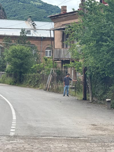 In Tumanyan, Armenia, a man in a dark grey t-shirt and blue jeans stands on a paved road, leaning on a vertical wooden pole. The road curves to the left in the foreground, marked with a white line and dashed white rectangles. Behind the man, a wire fence separates the road from overgrown vegetation. Further back, a multi-story building with light brown walls, a metal roof, and balconies is visible. Trees and foliage surround the building and fence. In the background, a lush, green mountainous landscape dominates the scene under a cloudy sky.
