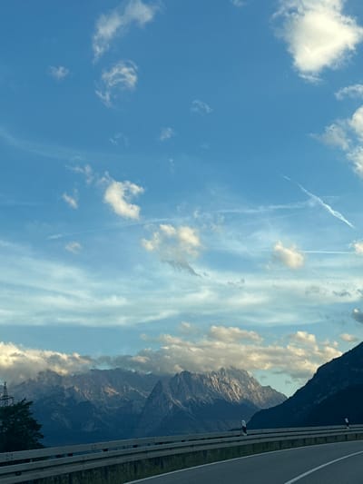 This image captures a scenic view of the Alps from a roadway in Farchant, Germany. The foreground features a paved road with white lane markings and a metal guardrail. Beyond the road, a line of mountains stretches across the horizon, their peaks illuminated by the sun. The mountains are partially obscured by clouds that hug their upper slopes. The sky is a vibrant blue, dotted with scattered white clouds, including some wispy contrails from aircraft. The lighting suggests it is either early morning or late afternoon. The overall scene is one of natural beauty and a sense of open space.