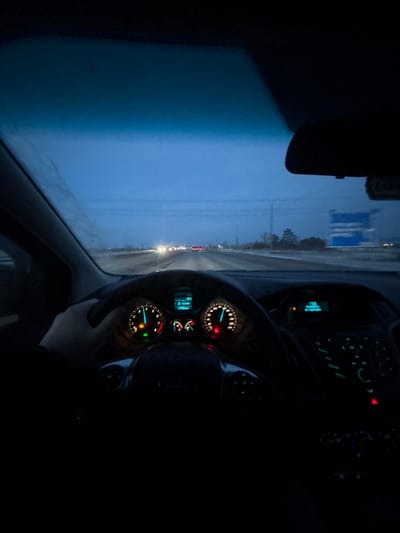 The image displays a first-person perspective from inside a moving vehicle on a road in Terent'yevo, Russia, during low-light conditions, indicative of dusk or dawn. A human left hand is positioned on the black steering wheel at approximately the 9 o'clock position. The vehicle's instrument cluster is illuminated: the speedometer indicates a speed between 100 and 110 km/h, with an active blue indicator light. The tachometer shows a green indicator light and a blue engine coolant temperature light. A central digital display within the cluster registers an ambient temperature of 3°C and a distance reading of 17336.0 km. The central console's infotainment system is partially visible to the right, illuminated with blue and green controls. The rearview mirror is visible at the top center. The windshield shows frost or condensation on its upper left portion and appears generally obscured by precipitation or residue. Outside the vehicle, a multi-lane road, appearing damp or lightly covered in snow/slush, stretches into the distance. Other vehicles are visible ahead, identifiable by their illuminated headlights and taillights. A large blue road sign with white, illegible text is present on the right side of the road. Utility poles or power lines line the roadside. The sky overhead is a uniform dark blue, suggesting an overcast environment.
