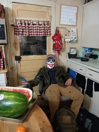 The image shows a person wearing a clown mask, a dark leather jacket, tan pants, and gray shoes, sitting in a cluttered room. The person is holding a blue smartphone in their left hand. Next to them on a table is a metal tray containing a watermelon (partially sliced) and an orange. Behind the person are kitchen cabinets and drawers, and a window with a red and white checkered curtain. A calendar and a red backpack are hanging on the wall. The exact location in Clarksville, United States is not discernible from the image.
