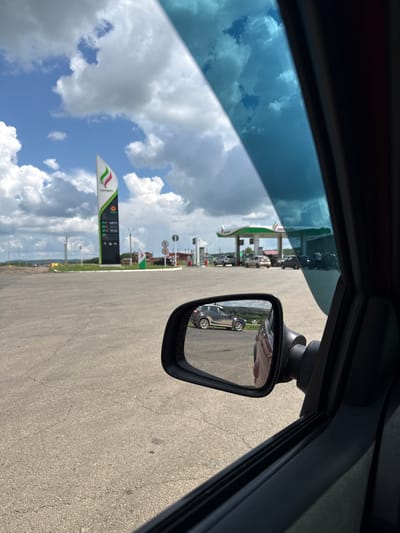 The image shows a view from inside a car looking out the window and in the side mirror, at a gas station. The gas station is identified as "GAZPROM" by signage. The scene appears to be in Kachimir, Russia, as indicated by the presence of Gazprom, a Russian energy company. The sky is cloudy with patches of blue. In the foreground, the side of the car and the side mirror are visible. The side mirror reflects other cars and the gas station canopy. In the background, the gas station has multiple pumps and a tall sign displaying prices. The overall environment suggests a typical day at a Russian gas station.