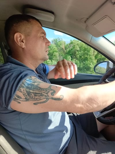 A man with a buzz cut is driving a car, seen from the chest up. He is wearing a dark blue polo shirt and dark shorts. He has a large, intricate tattoo of an octopus on his left arm, which is visible as he grips the steering wheel. His right hand is resting on the steering wheel, and his left arm is extended slightly. He appears to be looking straight ahead, with a neutral expression. The car's interior is visible, including the sun visor with "AIRBAG" printed on it and the side mirror reflecting a clear blue sky and green trees. The scene is set outdoors during the day, likely with sunny weather, given the bright light and clear sky. The background outside the car windows consists of lush green trees, suggesting a rural or suburban setting. The image was taken in Vyazovka, Russia.
