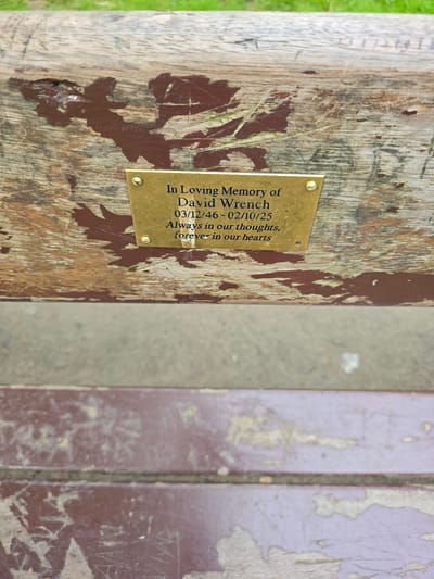A brass plaque is affixed to a weathered wooden surface, likely a park bench, with four visible rivets. The plaque bears an inscription in dark lettering. The inscription reads: "In Loving Memory of David Wrench 03/12/46 - 02/10/25 Always in our thoughts, forever in our hearts." The wooden surface exhibits peeling brown paint and has a rough texture. The background is blurred but suggests a grassy area. The image was taken outdoors, likely in a public space. The location in the city of Stockton-on-Tees, United Kingdom is not discernible from the image itself.