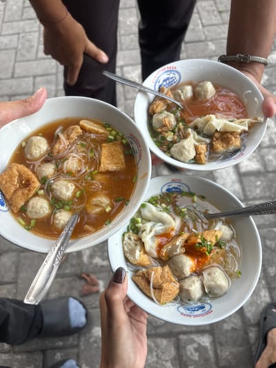 Three bowls of bakso, a traditional Indonesian meatball soup, are being held by individuals outdoors, likely on a sidewalk given the cobblestone-like ground visible. The bowls contain meatballs, noodles, fried tofu, and chopped scallions, with one bowl also featuring dumplings. The lighting suggests daytime. The image does not provide enough visual information to confirm the specific location in Kalijaten, Indonesia.