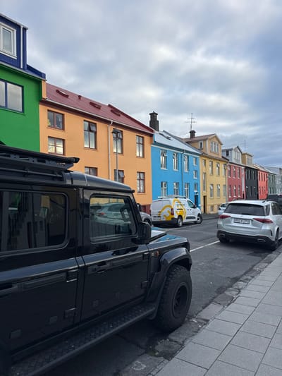 The image shows a street in Reykjavik, Iceland, lined with brightly colored buildings. These buildings are painted in a variety of colors, including blue, green, yellow, and orange. There are parked cars along the street. The sky is overcast with a layer of clouds. The perspective is from a low angle, emphasizing the height of a black SUV on the side of the road.