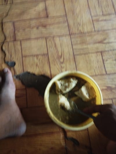 A person's bare foot is visible in the lower left corner of the image. The foot is partially obscured by a crack in the wooden floor. A yellow bowl containing a brown liquid and pieces of light-colored food is in the lower right corner. A hand is visible holding a spoon that is partially submerged in the bowl. The coordinates 10.852, 11.426 correspond to a location in Bajoga, Nigeria.  The scene is a close-up of someone eating a meal on a wooden floor. The meal appears to be a type of soup or stew.  The lighting is dim. The environment is domestic.