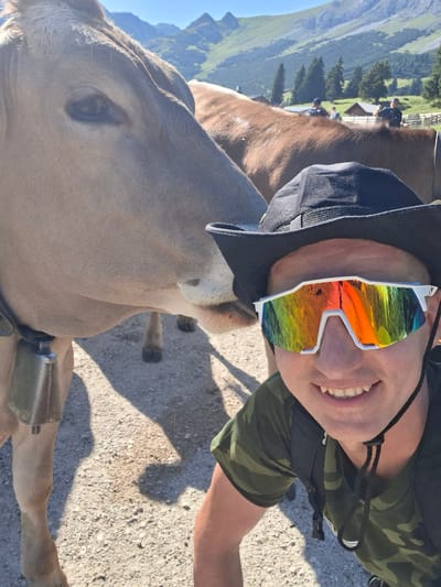 A man wearing a camouflage shirt, black hat, and white-framed sunglasses with rainbow-colored lenses smiles at the camera. He is in the foreground of the image, and a light brown cow is to his left, its head close to his face. The cow has a bell around its neck. In the background, other cows are visible, as well as a green, mountainous landscape with trees and a small building. The ground is covered in gravel.