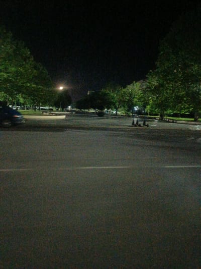 The image depicts a street scene at night in Pivdenne, Ukraine. In the foreground, a paved road with white lane markings is visible. The middle ground shows a sidewalk area with trees lining the background, illuminated by artificial lighting. A white-colored planter or curb is present. A parked car is partially visible on the left side of the image. In the distance, several electric scooters are parked in a row. The sky is dark, indicating nighttime. The image is grainy due to low light conditions.