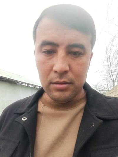 The image displays a head-and-chest portrait of an adult male. The subject has short, dark hair parted on the left, a medium complexion, and visible facial stubble. His gaze is directed downwards, resulting in a neutral expression. He is dressed in a dark, collared jacket worn over a light brown, ribbed crew-neck shirt. A silver-colored snap button is visible on the left lapel of the jacket. The background features an overcast sky. To the left, a light-colored building with a green corrugated metal roof is partially visible. To the right, bare tree branches are present against the bright sky, above a section of a light-colored wall or fence. The scene is located in Pichakchi, Uzbekistan.