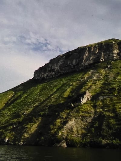 The image shows a steep, grassy mountainside rising from a dark body of water. The mountain has a rugged, rocky summit. Shadows are cast across the grassy slope, indicating it is daytime and the sun is at an angle. The sky above is partly cloudy and appears overcast. The scene is natural and undeveloped, with no signs of human structures or activity. The provided information about the location in Dubno, Ukraine, is not visually verifiable from the image itself.